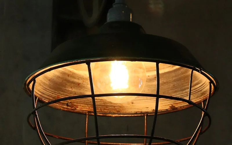 Pendelleuchte mit Drahtkorb und leuchtender Glühlampe vor dunkler Wand